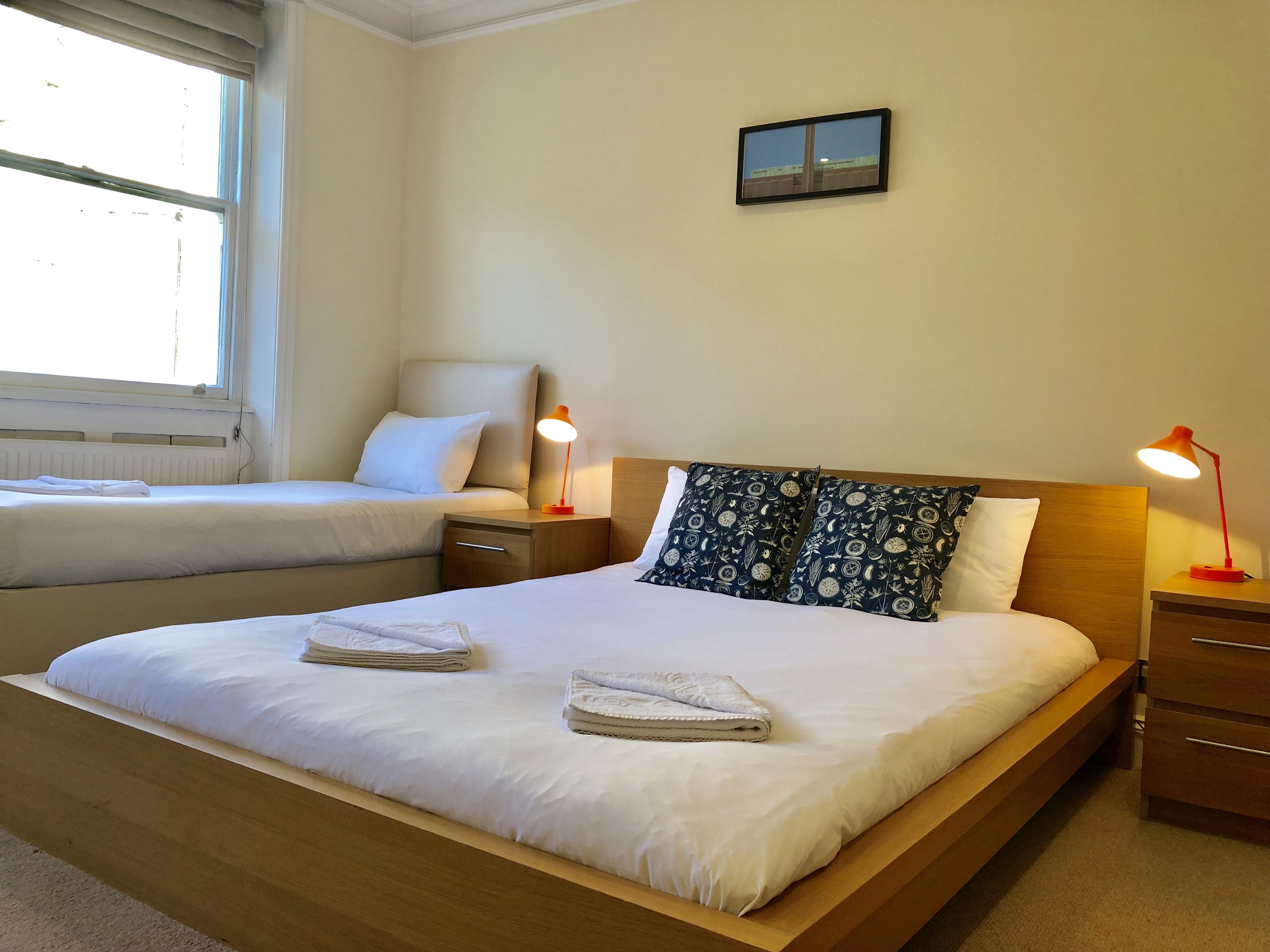 A hotel room with a large bed, a smaller bed, two bedside tables with lamps, a window, and a wall-mounted picture.