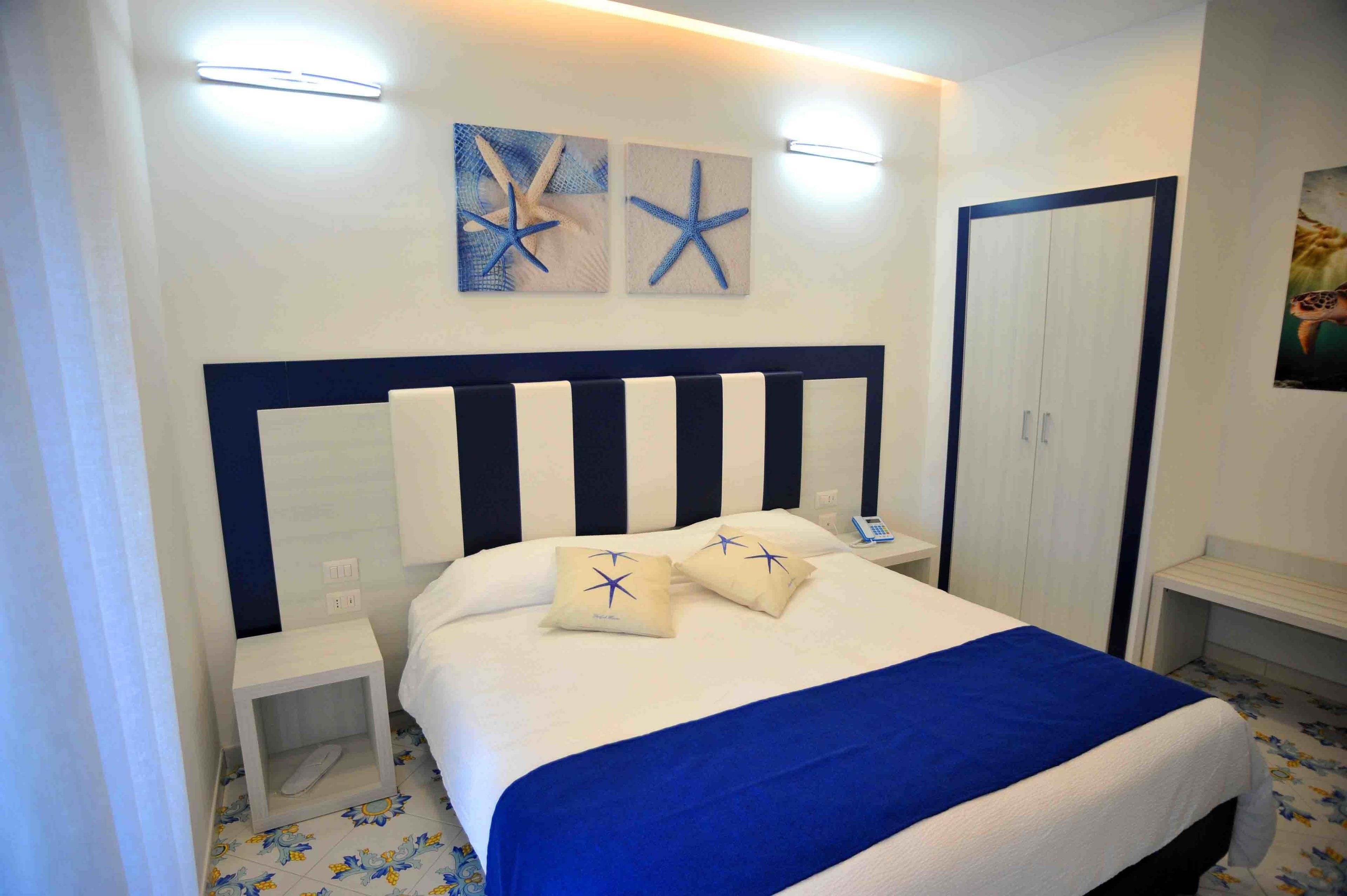 Un lit bien fait, avec une tête de lit à rayures bleues et blanches, deux coussins décorés d’étoiles de mer et un plaid bleu.