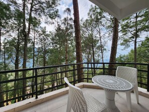 Un balcón con una mesa y sillas blancas, con vistas a un bosque de árboles altos y un cuerpo de agua.