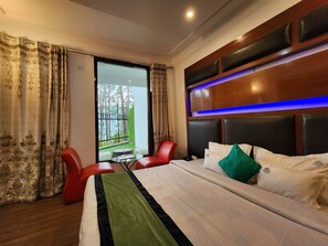 Hotelkamer met een groot bed, een rode stoel, een glazen tafel en uitzicht op een balkon met hekwerk en groen.