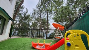 A playground with a red slide and a climbing structure, surrounded by a green fence and tall trees. - Hotel Bliss (Kasauli)