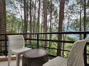 Een balkon met twee witte rieten stoelen en een ronde rieten tafel, met uitzicht op een bebost gebied.