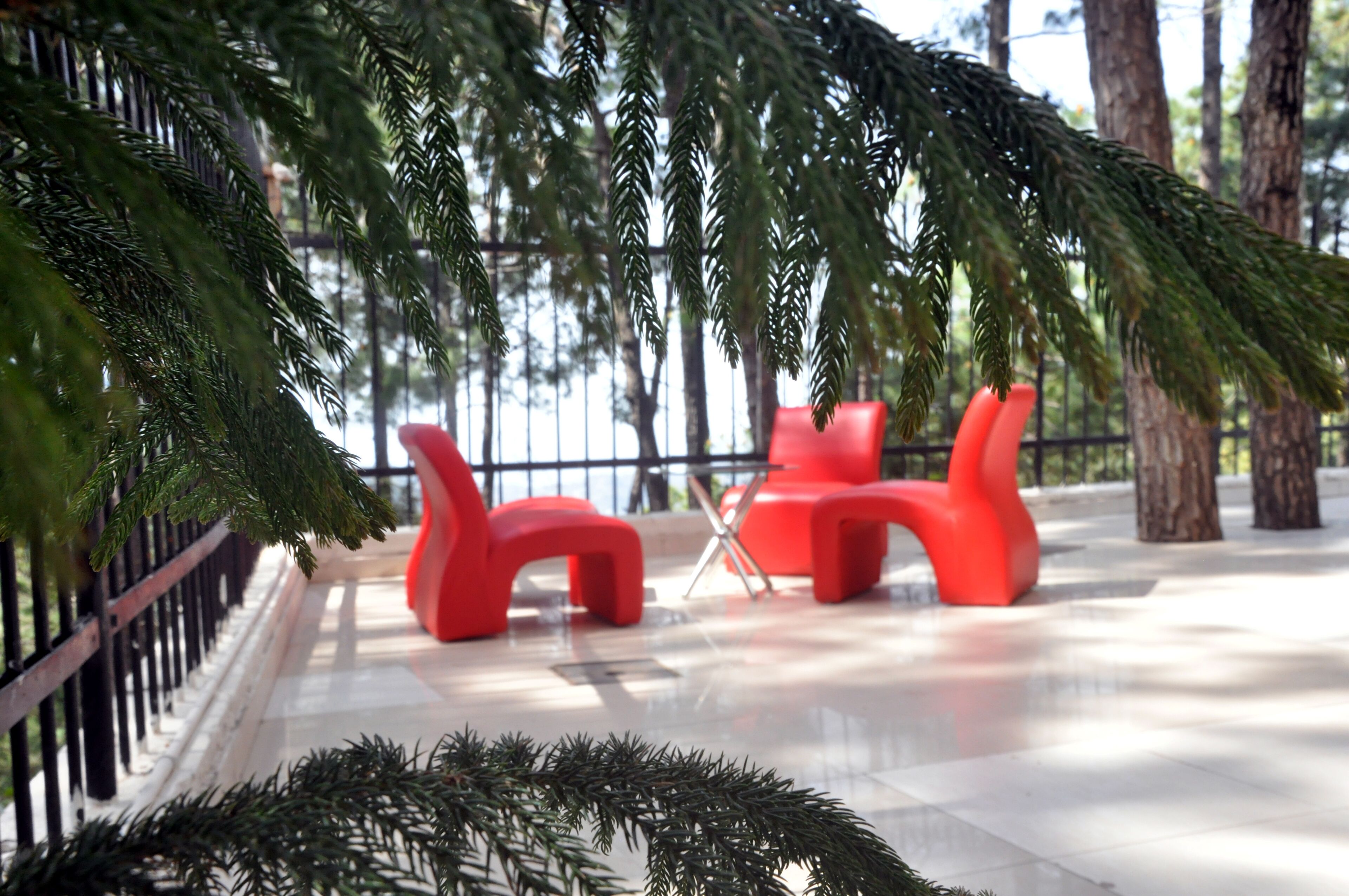 A modern outdoor seating area with red chairs and a glass table, surrounded by greenery and a metal fence.