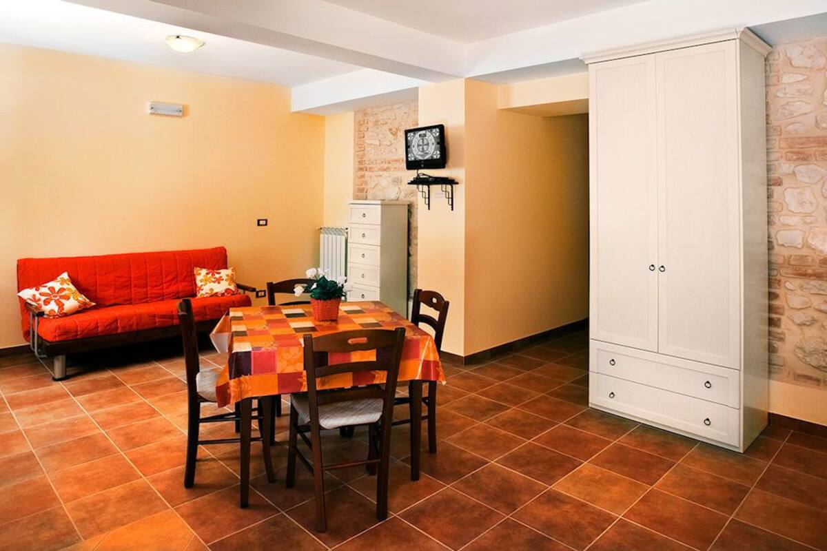 Ein Essbereich mit kariertem Tischlaken, Holzsesseln, einer roten Couch mit gemusterten Kissen, einem weißen Schrank und einer Steinwand.