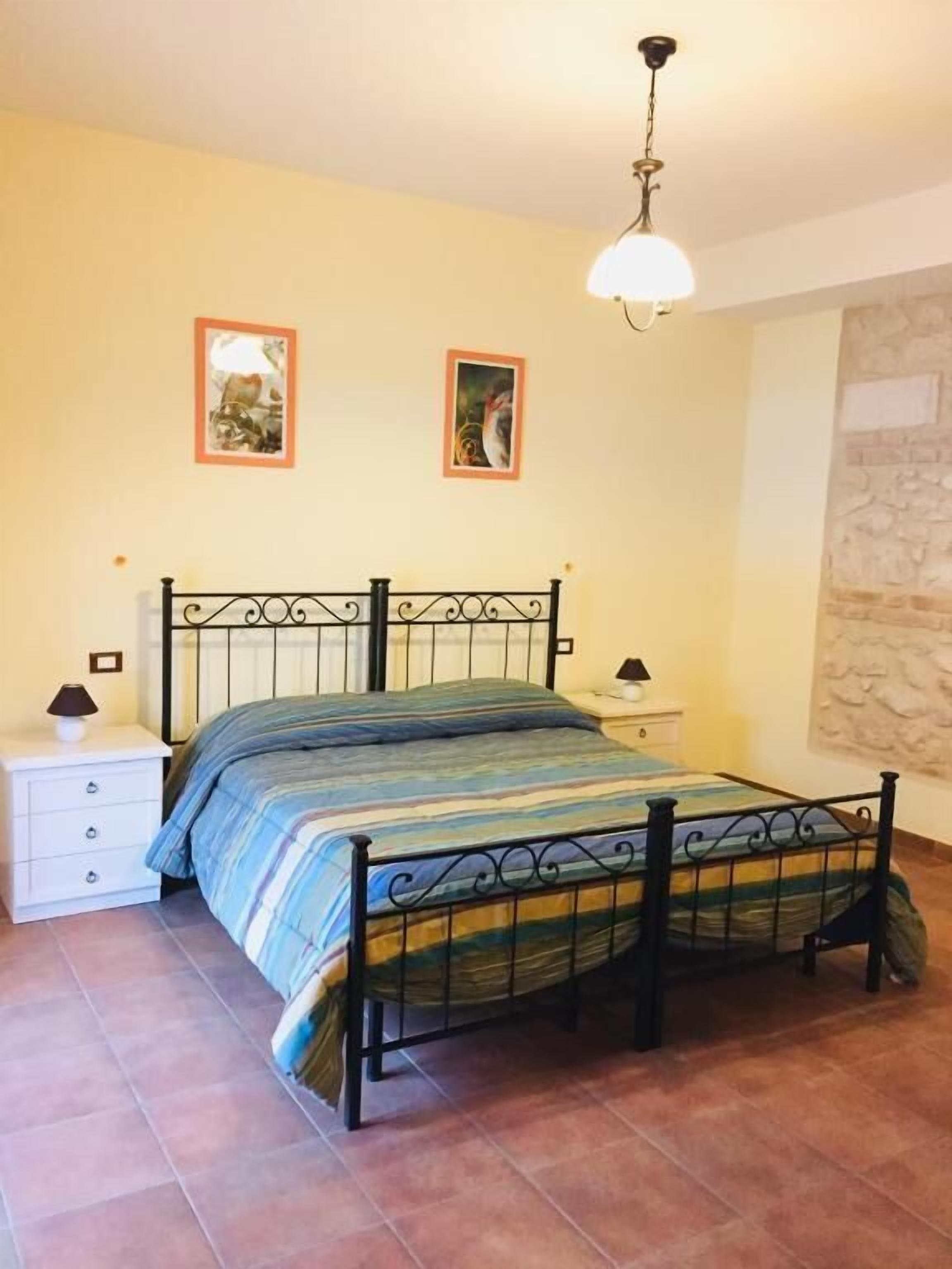 Un dormitorio con estructura metálica para la cama, una colcha a rayas, dos mesitas de noche con lámparas, y dos cuadros en la pared.