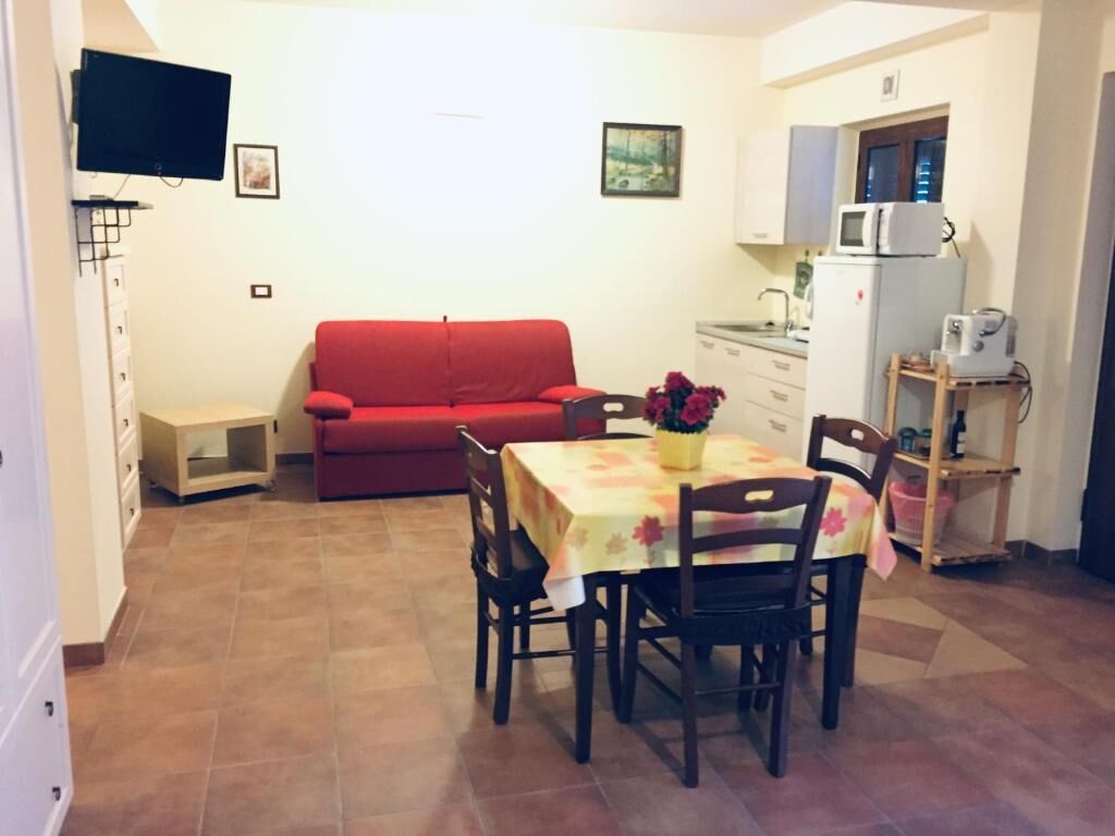 Un espacio habitable compacto con un sofá rojo, una mesa de comedor con sillas, una cocina con microondas y refrigerador, y un televisor montado en la pared.