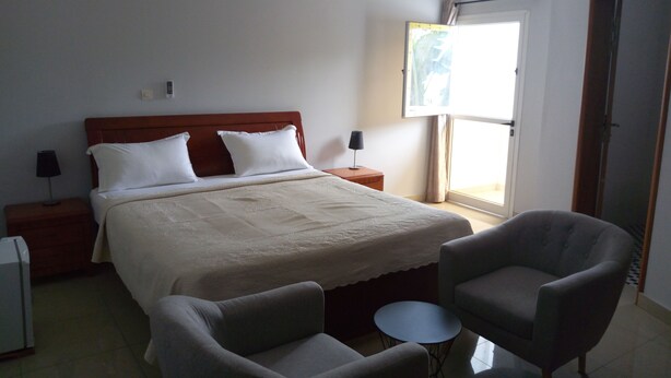 Una habitación de hotel con cama, dos mesitas de noche, una lámpara, una silla, una mesita redonda y una ventana con cortinas.