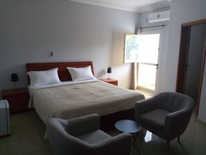 A hotel room with a bed, two bedside tables, a lamp, a chair, a small round table, and a window with curtains. - Hotel Edenia (Libreville)