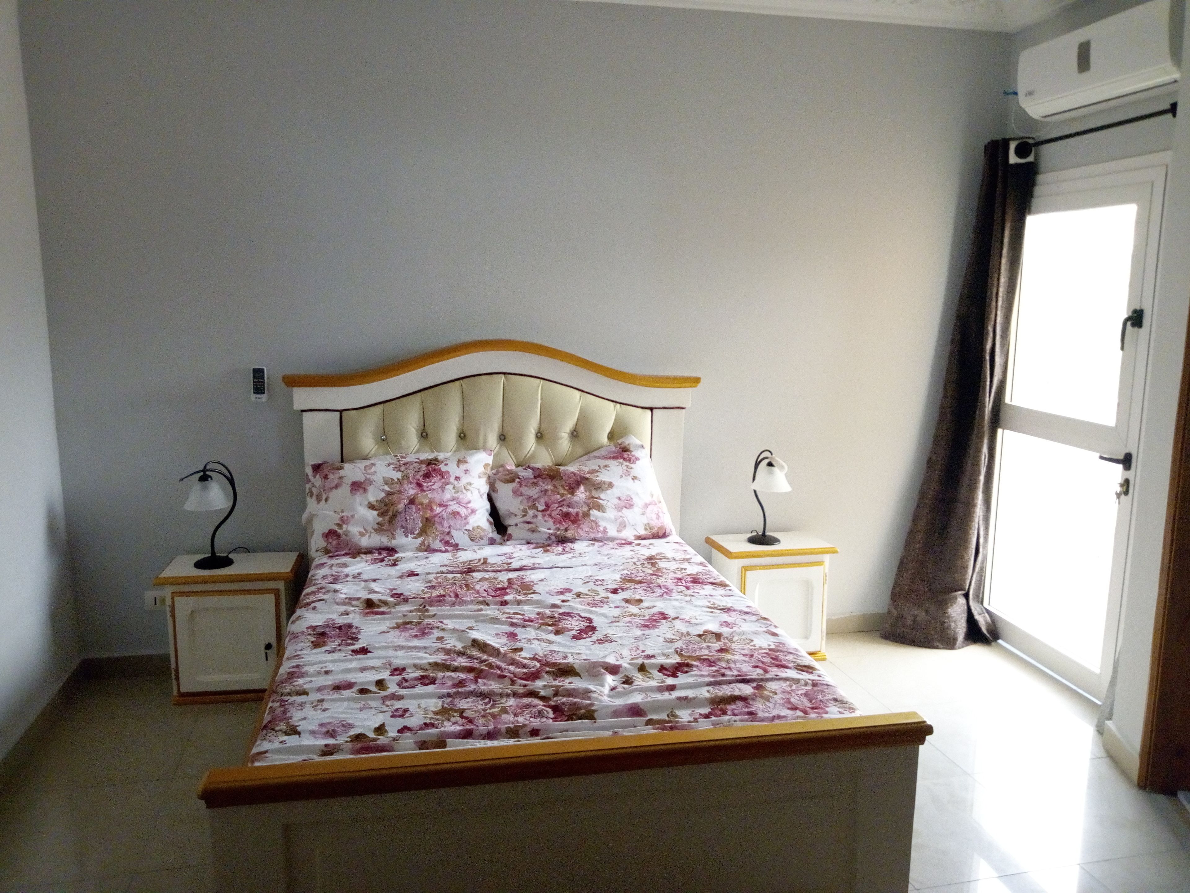 Une chambre à coucher comprenant un lit, deux tables de chevet, une commode, une fenêtre avec des rideaux et un climatiseur fixé au mur.