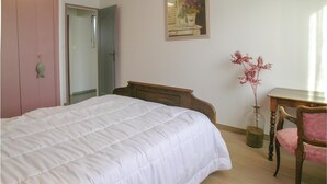 Ein Schlafzimmer mit einem Bett, einem Nachttisch aus Holz, einem Stuhl, einer Blumenvase und einem Spiegel an der Wand.