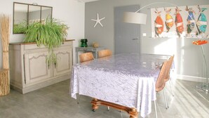 Ein Essbereich mit einem Tisch, der mit einer Abdeckung geschützt ist, einem Holzstuhl, einem Schrank mit Pflanzen, einer Meerstern-Dekoration und einem Fischgemälde an der Wand.