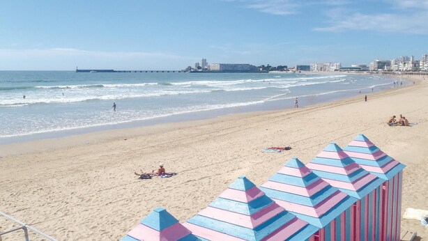 Ein Strand mit einem Pier im Hintergrund, einem sandigen Ufer und einem farbenfrohen Gebäude im Vordergrund.
