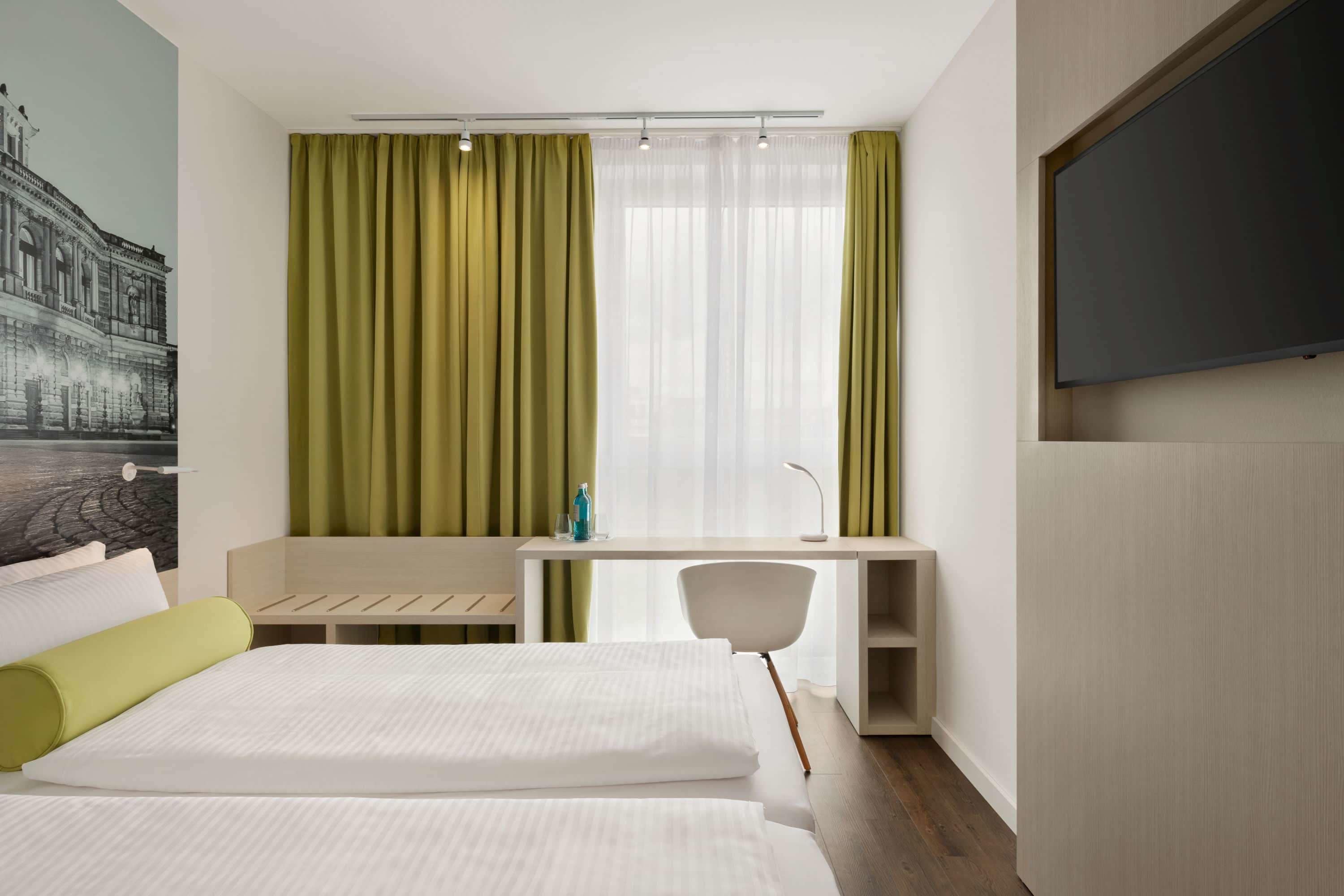Habitación de hotel con dos camas, un escritorio, una silla, un televisor y un ventanal con cortinas.