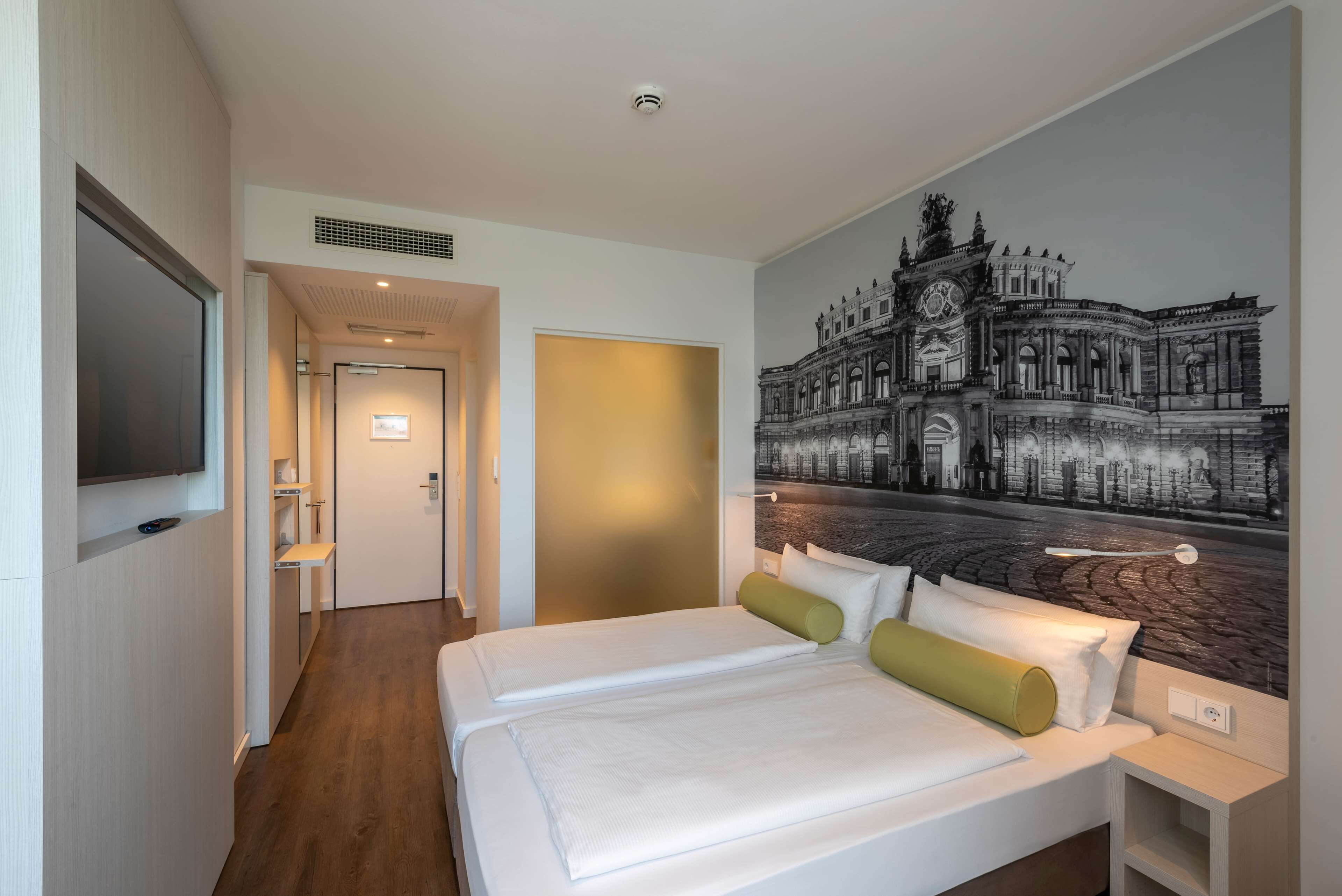A hotel room with a large bed, a flat-screen TV mounted on the wall, and a mural of a historic building.