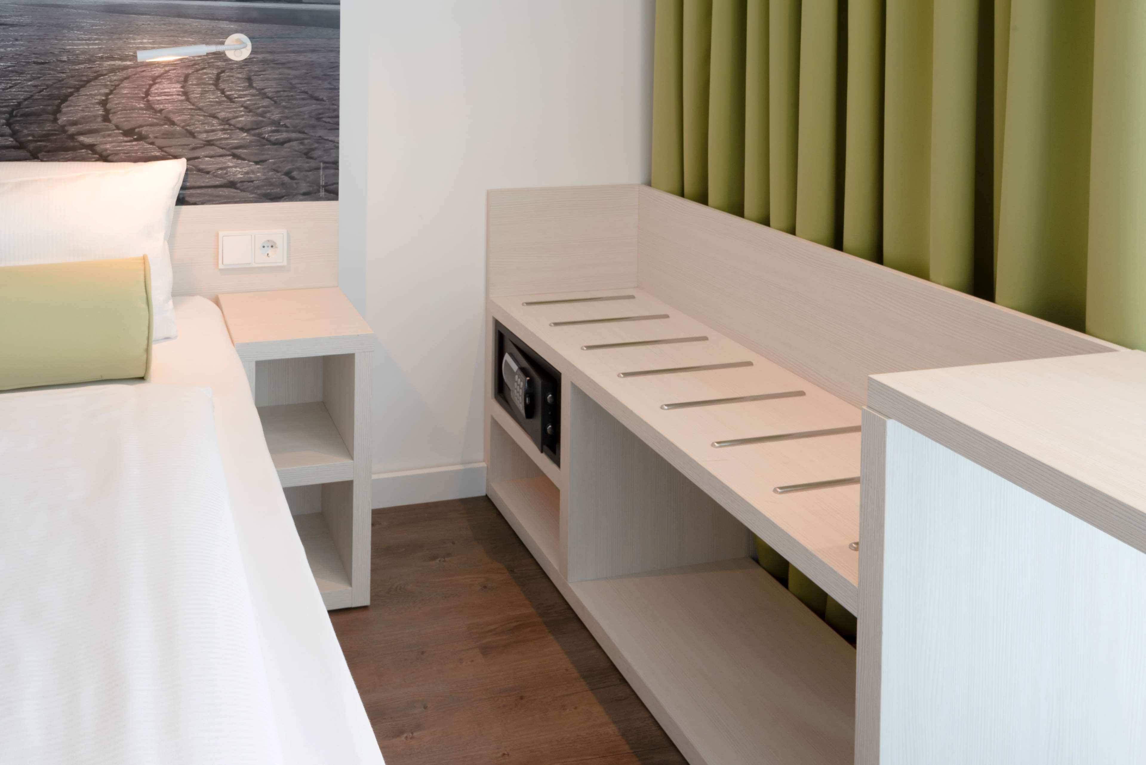 Una habitación de hotel moderna con una cama blanca, una mesita de noche pequeña, un estante empotrado con ganchos, vista a una calle empedrada y un panel de pared verde.