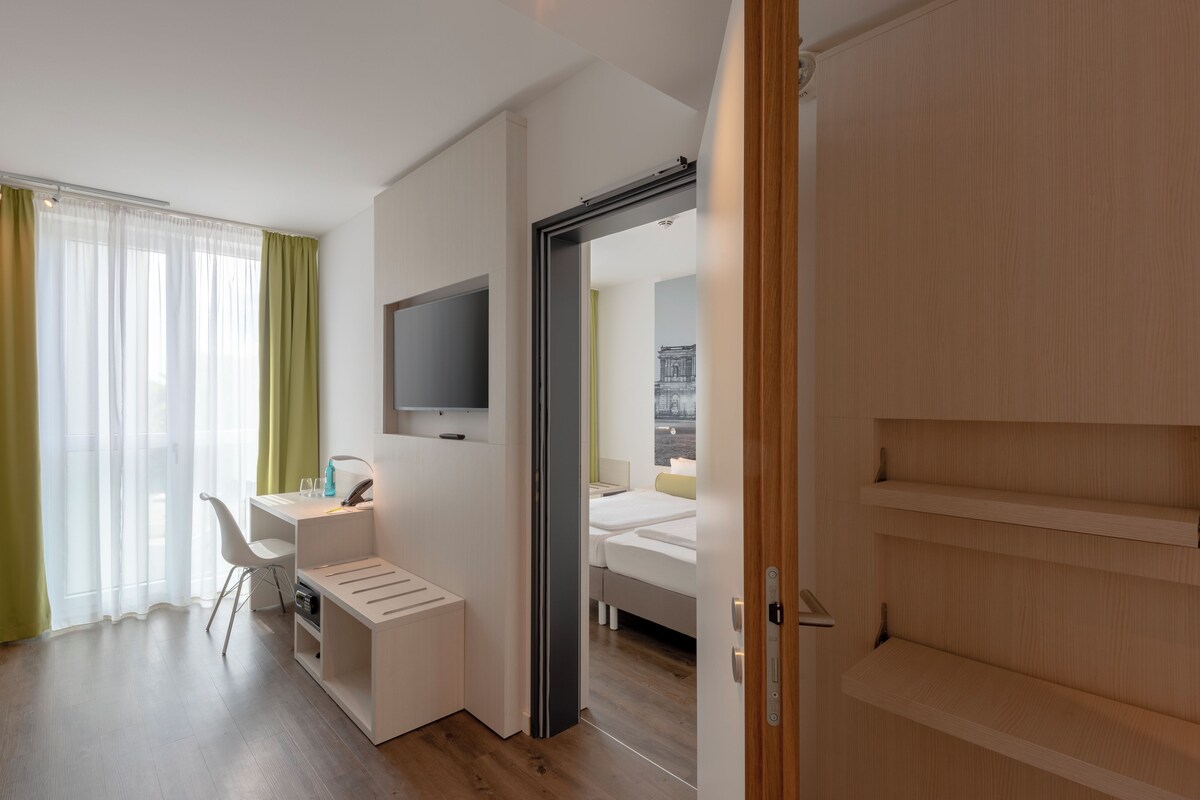 A hotel room with a bed, a desk with a chair, a television, and a view of a building through the window.