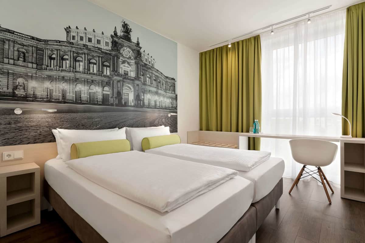 Habitación de hotel con dos camas, un escritorio, una silla y un mural grande de un edificio histórico.