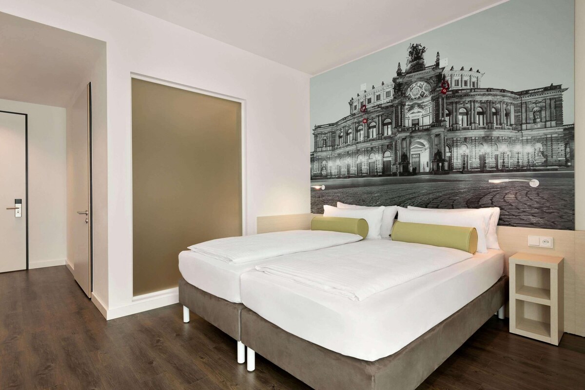 A double bed with white bedding and two pillows, a nightstand, a wall-mounted clock, and a large black-and-white cityscape painting.