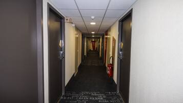 A hotel hallway with a carpet featuring text in multiple languages, doors on both sides, and a fire extinguisher on the right.