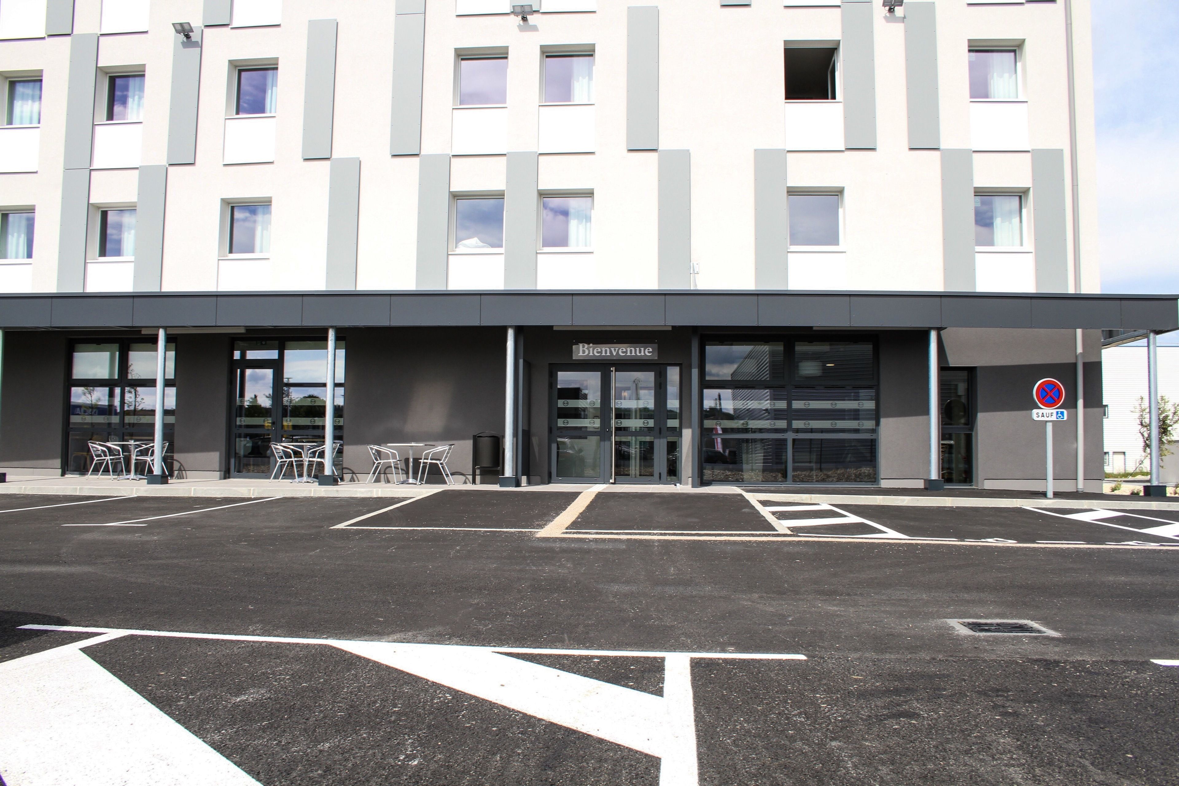 A modern building with a 'Bienvenue' sign, a parking lot with marked spaces, and a 'No Parking' sign.
