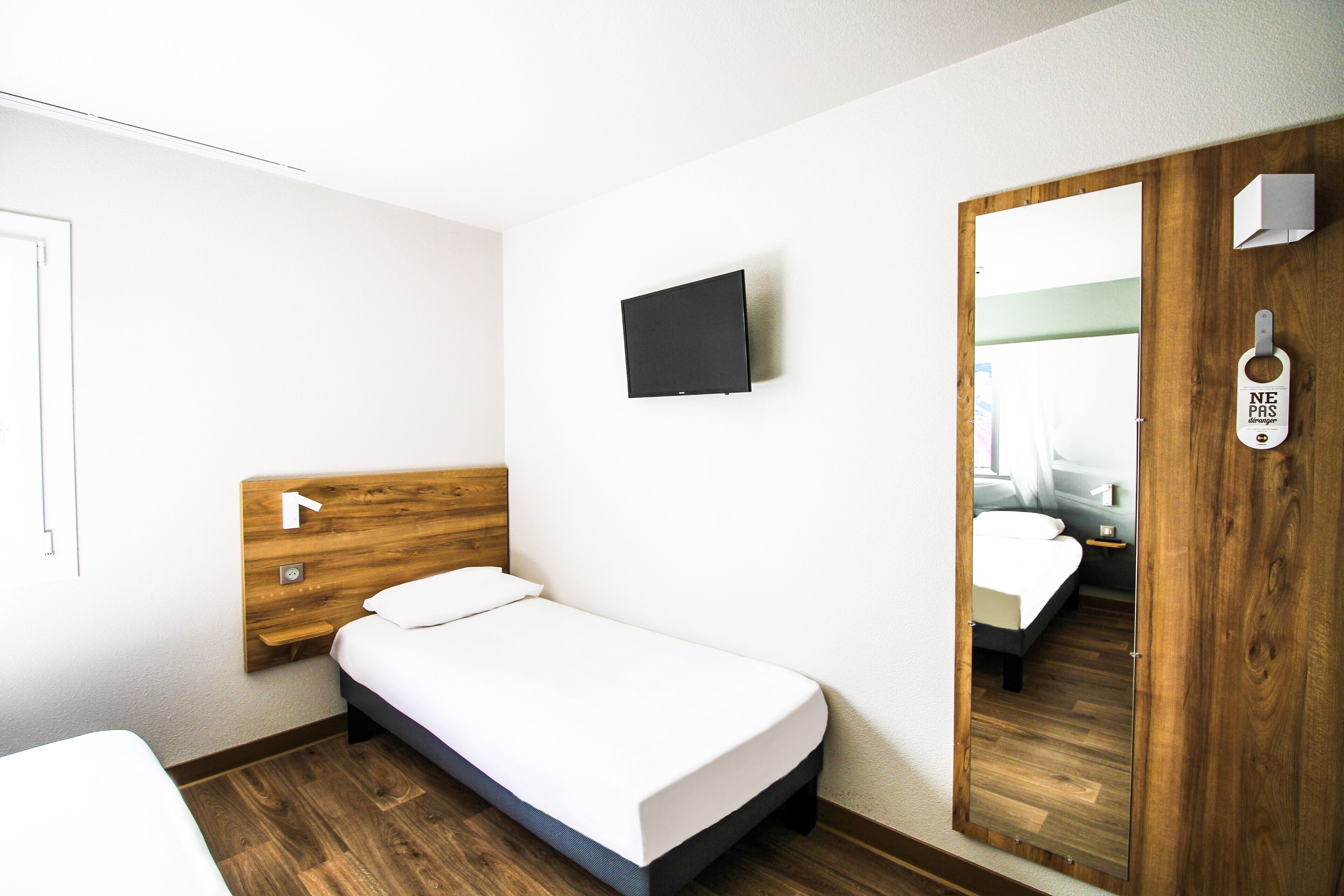 Un dormitorio pequeño y ordenado con una cama individual, un televisor montado, un cabecero de madera y un espejo en la puerta.