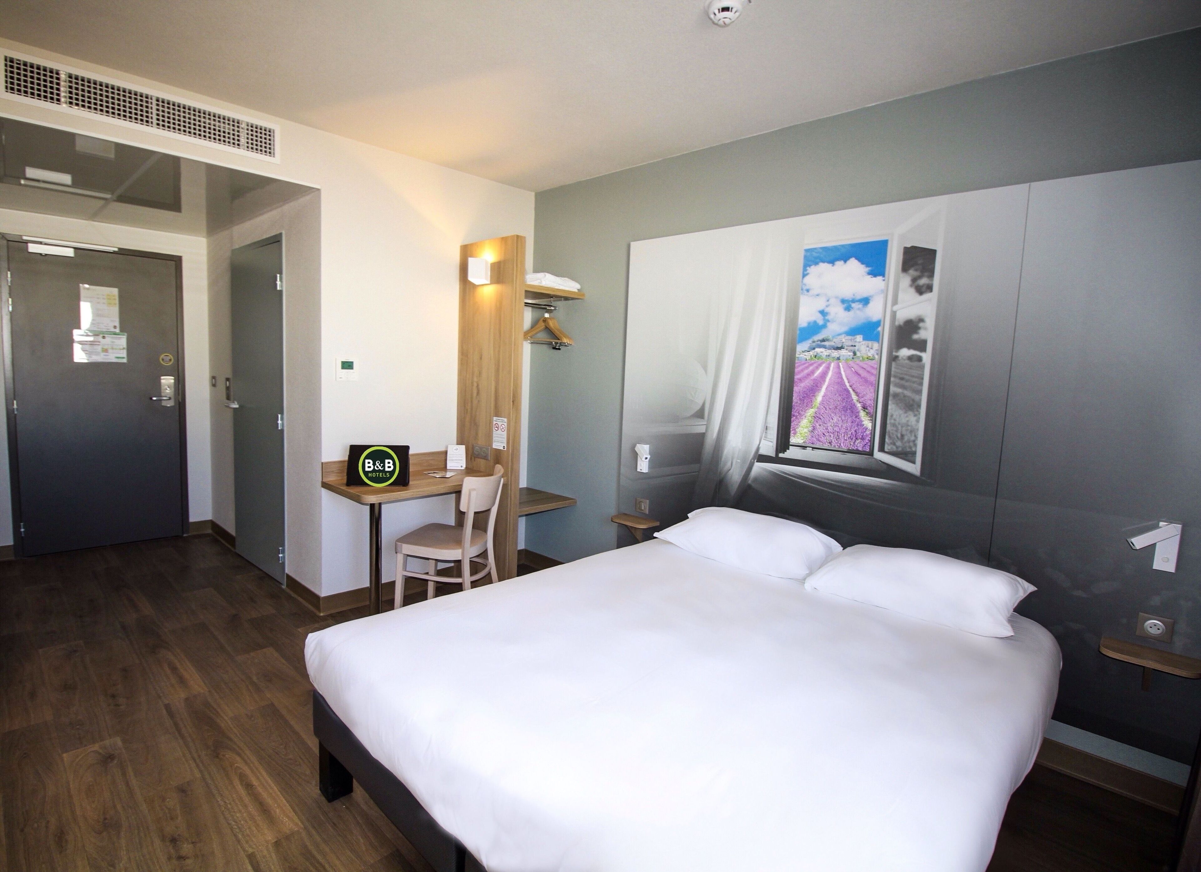 Habitación de hotel con cama, escritorio con silla, ventana con vistas a campos de lavanda y una puerta.