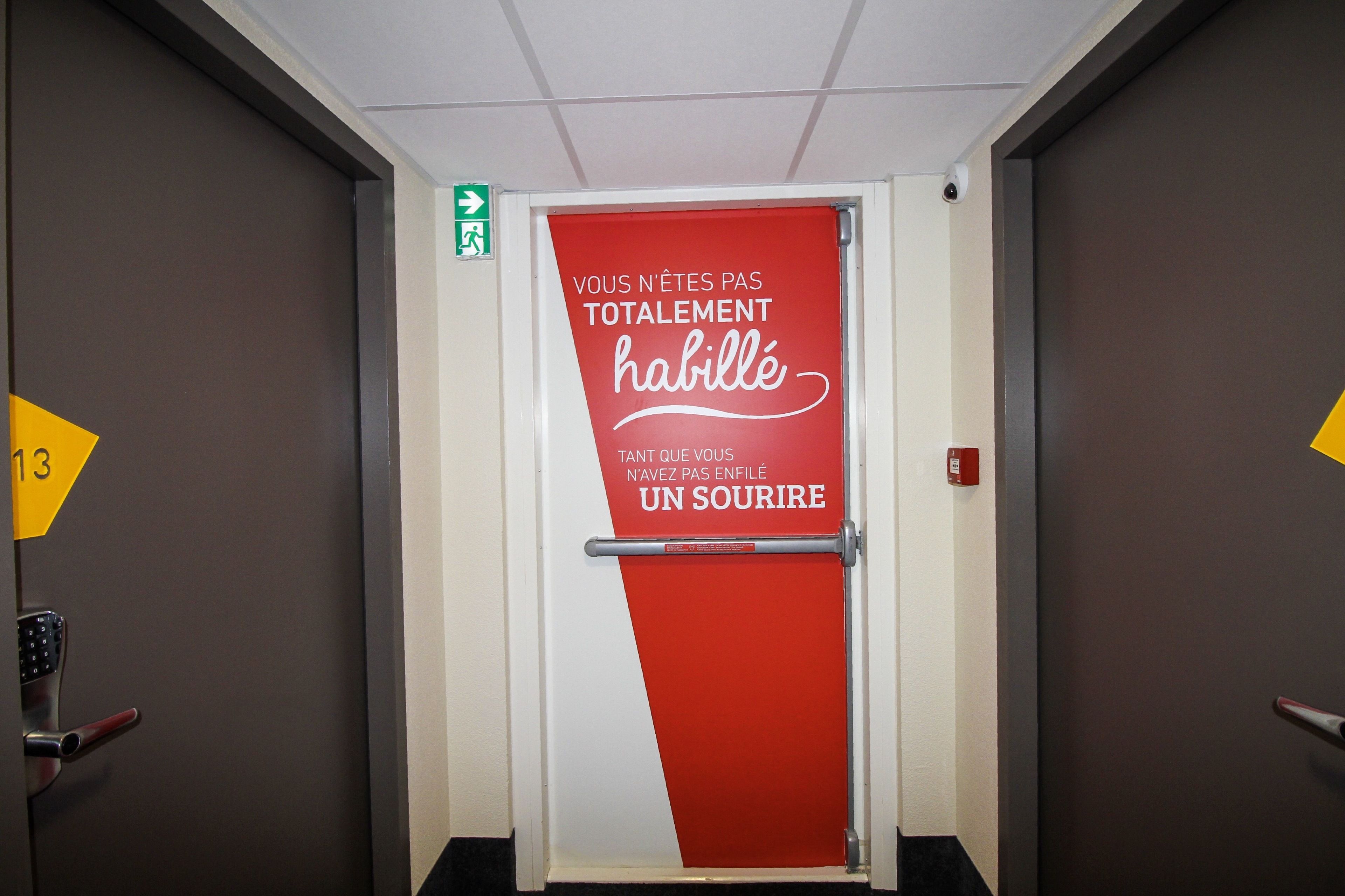 Puerta de habitación de hotel con un cartel rojo y blanco que dice "VOUS N'ÊTES PAS TOTALEMENT habillé" y "TANT QUE VOUS N'AVEZ PAS ENFILÉ UN SOURIRE".