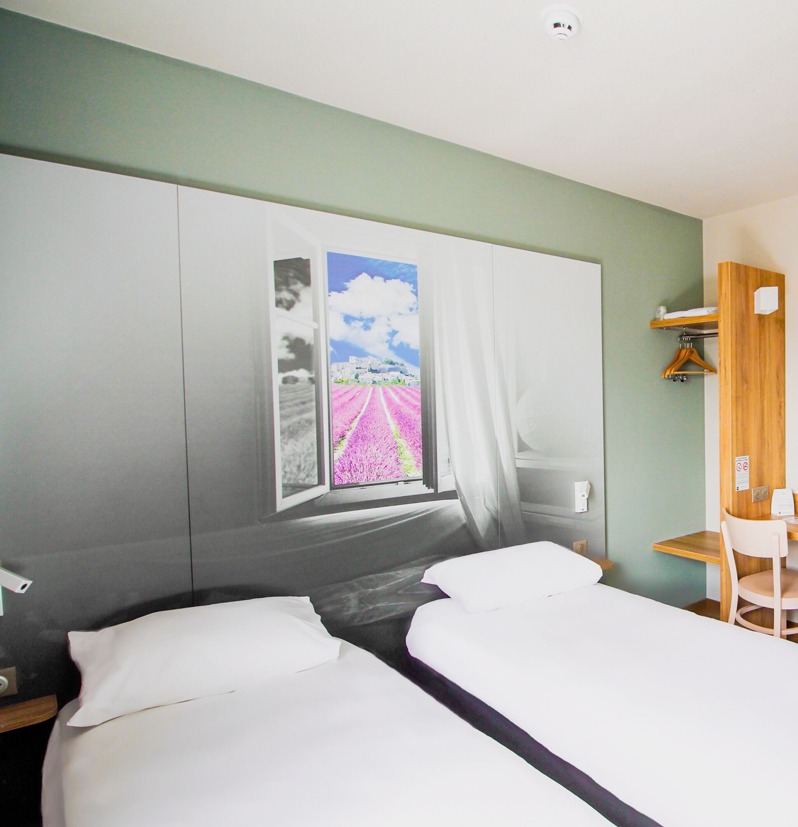 Quarto de hotel com uma cama, uma escrivaninha e uma cadeira. Há uma janela com cortinas e um quadro na parede.