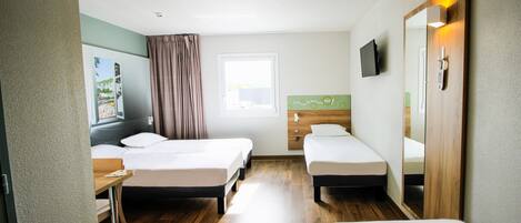 Habitación de hotel con dos camas individuales, un escritorio, una silla, un ventanal con cortinas, un televisor de pantalla plana montado en la pared y una puerta de madera con espejo.