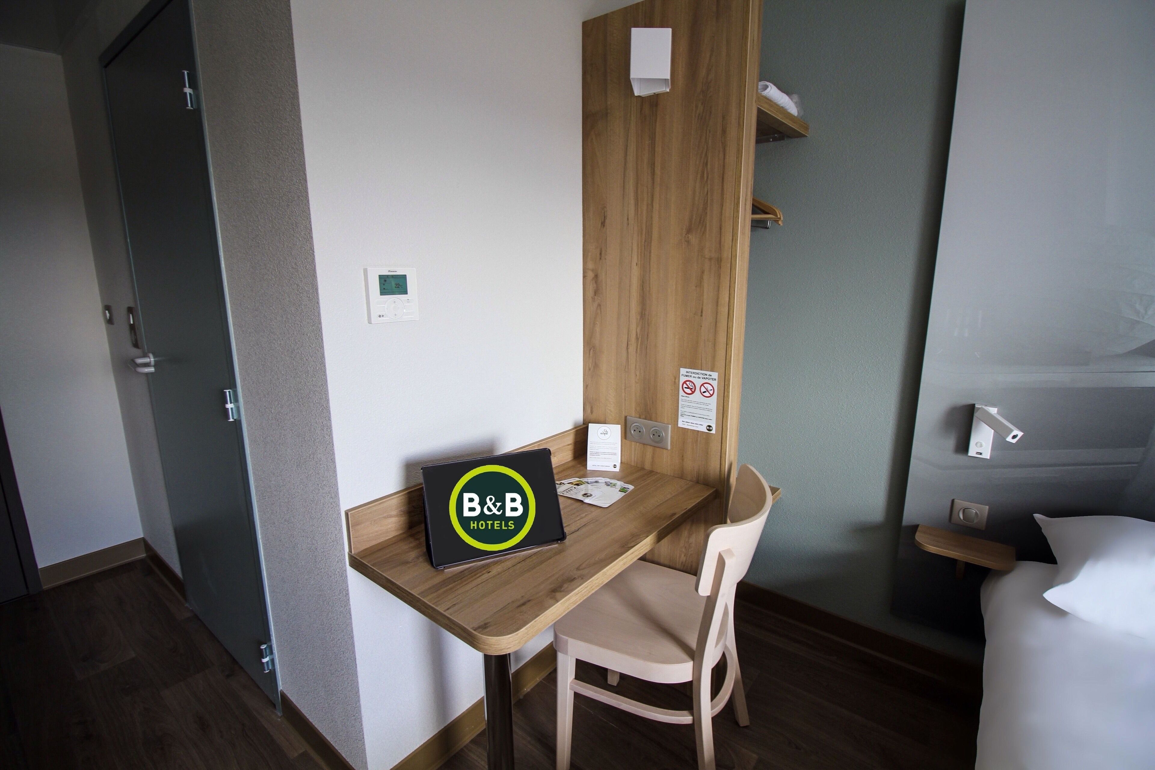 A hotel room with a wooden desk, a laptop with 'B&B HOTELS' logo, a bed, and a bedside table.