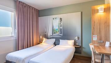 Ein Hotelzimmer mit zwei Einzelbetten, einem Holztisch, einem Stuhl, einem Fenster mit Vorhängen und einer Wandleuchte.