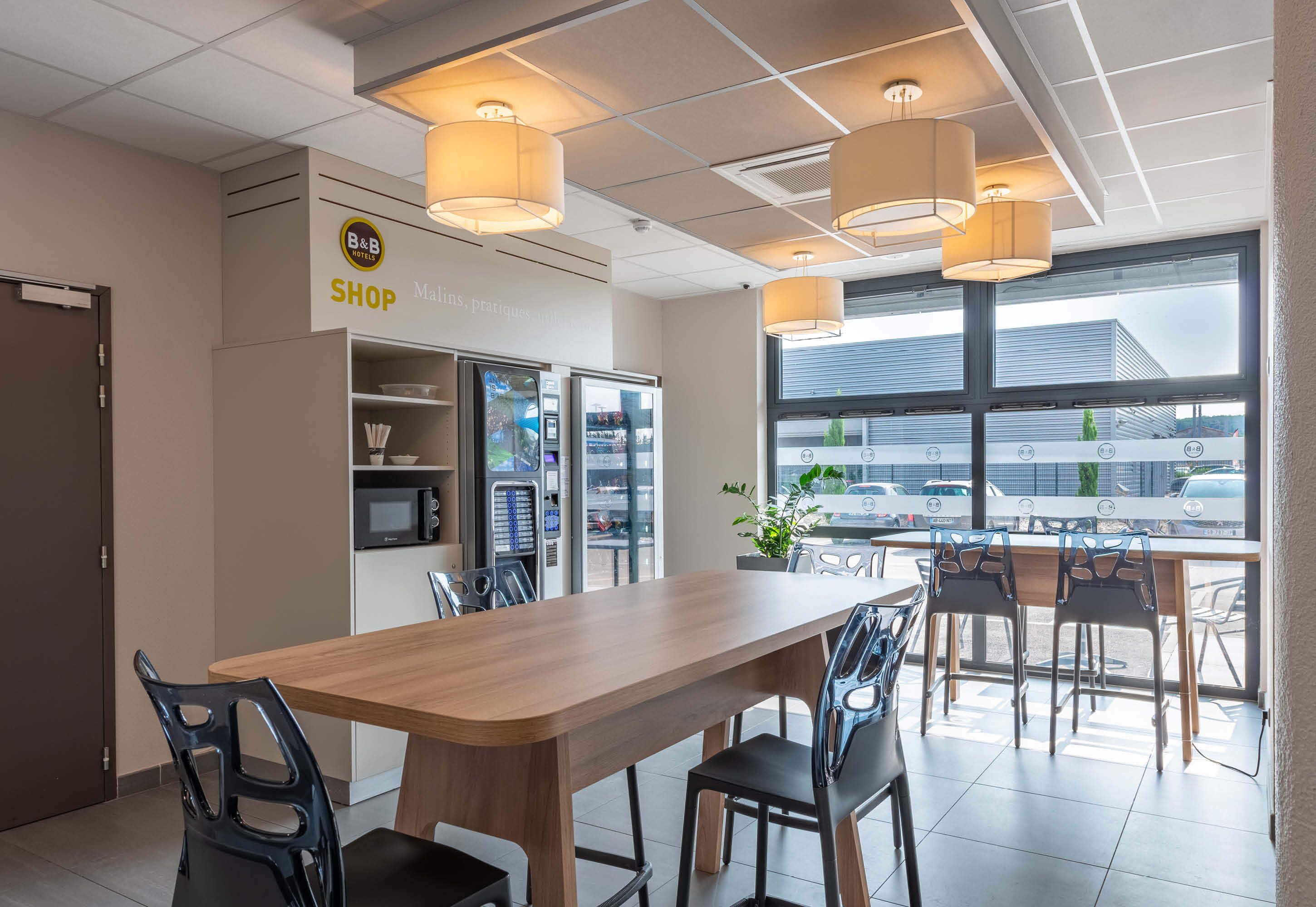 Un comedor moderno con una mesa y sillas de madera, un letrero de tienda de B&B, máquinas expendedoras y vistas a través de amplios ventanales.