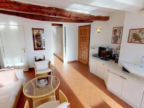 Un espace de vie compact comprenant un coin cuisine, un coin repas avec une table en verre et un canapé. On y trouve une porte en bois et un luminaire fixé au mur.