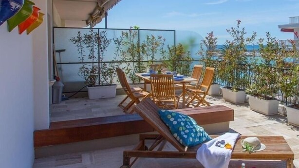 Eine sonnendurchflutete Terrasse im Freien mit einer Holzliege, einem Esstisch mit Stühlen und Topfpflanzen.