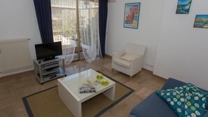 Ein Wohnzimmer mit Fernseher, Couchtisch, Sofa, Sessel und einem Fenster mit Vorhängen.