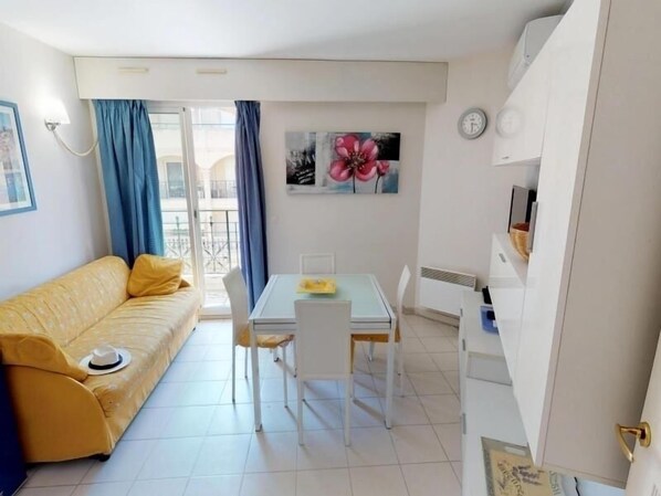 A compact living space with a yellow sofa, a dining table with chairs, a painting on the wall, and a white cabinet.