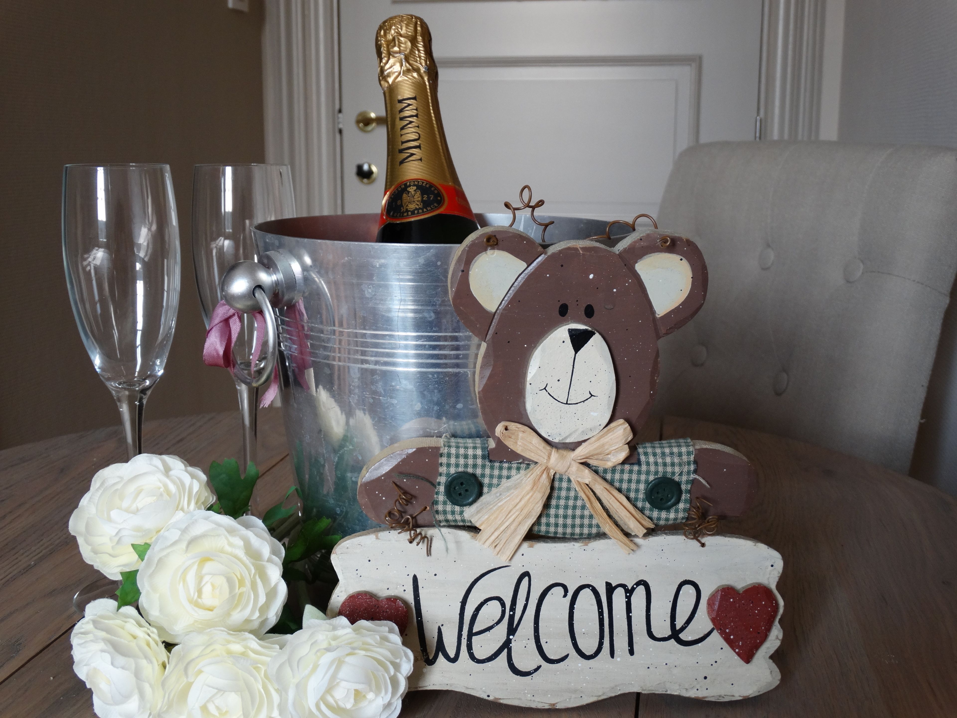 A champagne bottle in a metal bucket, two champagne flutes, a teddy bear, and a welcome sign with roses.