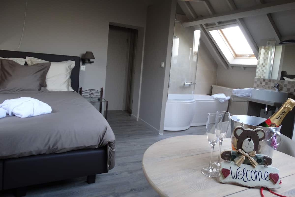 A bedroom with a bed, bedside table, and a small round table with a bottle of champagne, glasses, and a teddy bear decoration.