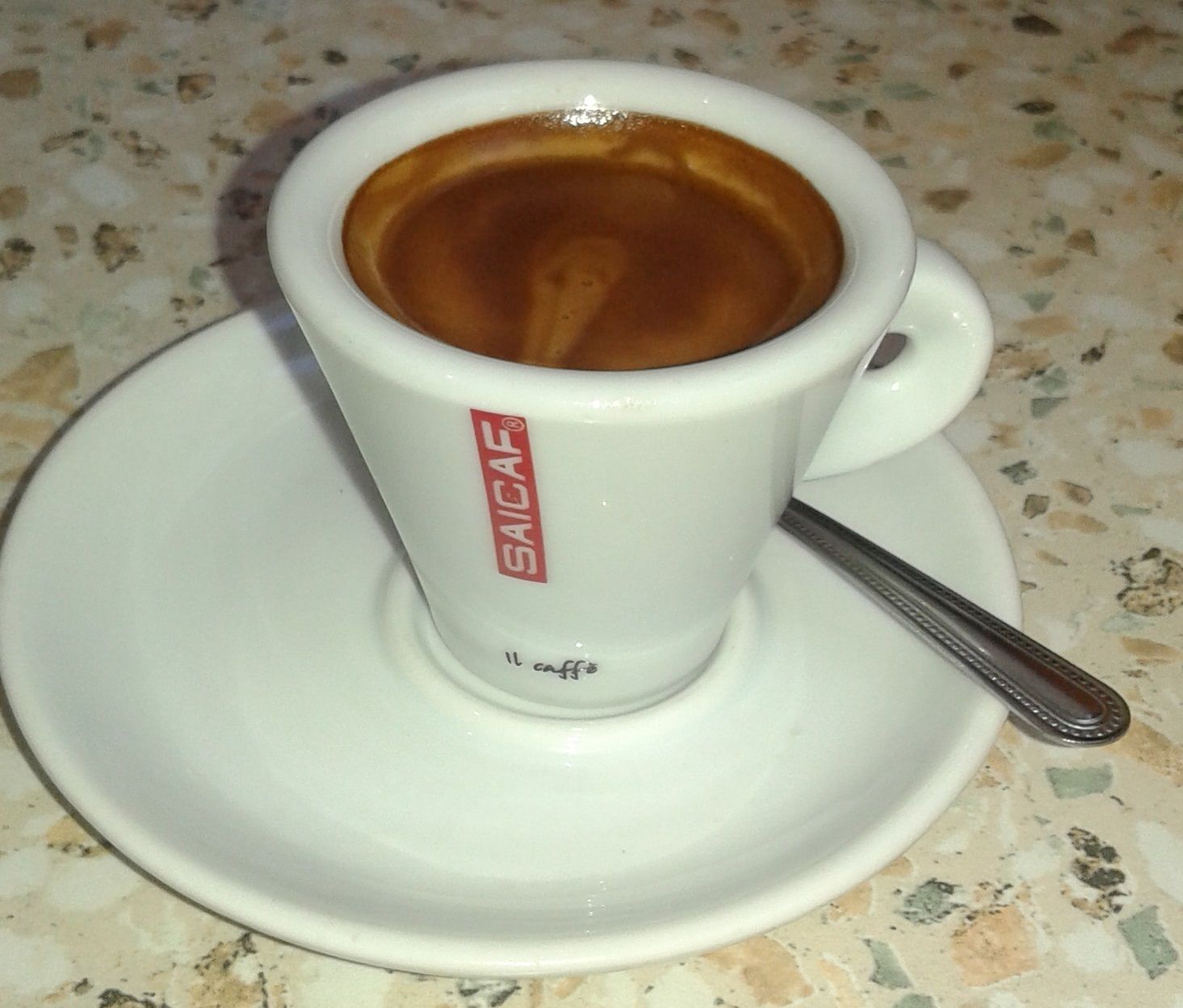 Een kop koffie op een schotel, met een lepel, op een gevlekt aanrecht.