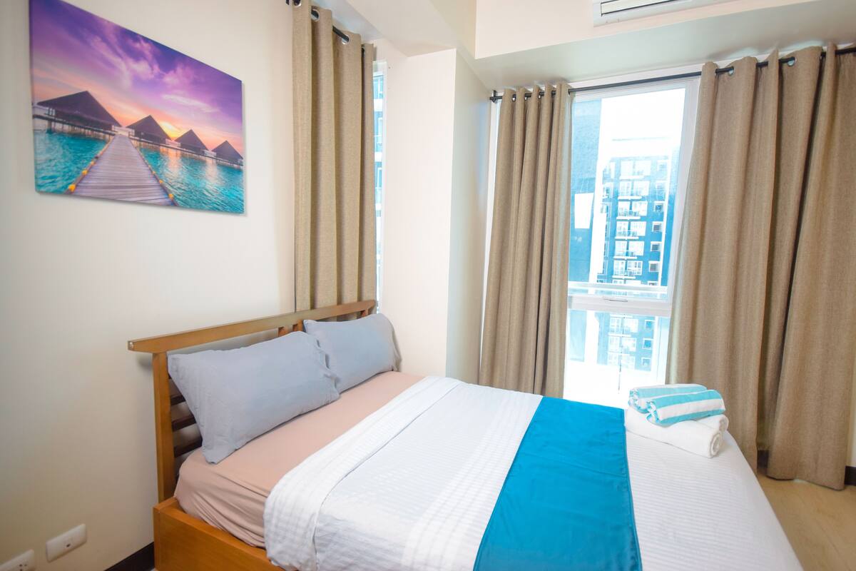 A hotel room with a bed, a painting of a sunset over a pier, and a window with curtains.