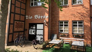 Ein Backsteinhaus mit einem Schild, auf dem "Die Pferde" steht. Vor dem Haus sind Fahrräder abgestellt, ein Tisch und Stühle stehen bereit, und es gibt eine grüne Außenfläche.