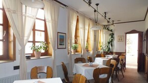 Ein Essbereich mit Tischen und Stühlen, großen Fenstern mit Vorhängen, Topfpflanzen und einer Kristallleuchtre.