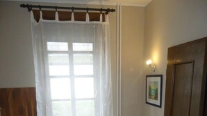 Ein kleiner, gut beleuchteter Raum mit einem Bett, einem Nachttisch, einem Holzschrank und einem Fenster mit durchsichtigen Vorhängen.