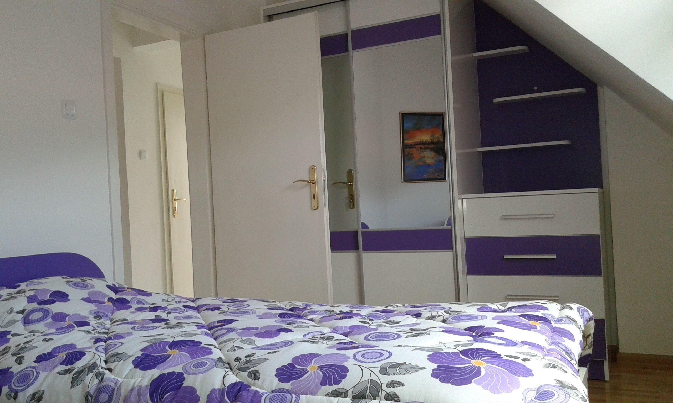 A bedroom with a bed, a wardrobe with a purple accent, and a painting on the wall.
