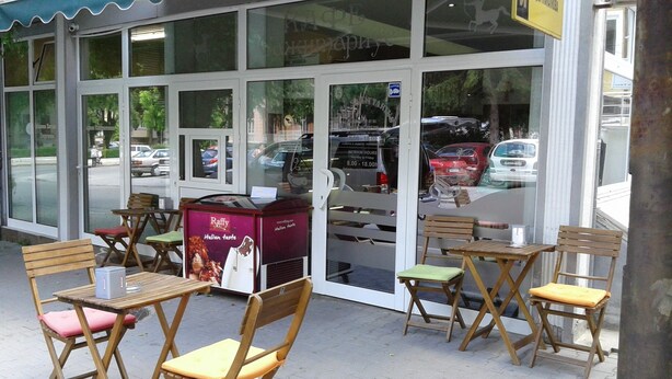 An outdoor seating area with wooden tables and chairs, a vending machine, and a glass storefront with reflections of cars and trees. - Sagittarius Apartment (Kyustendil)