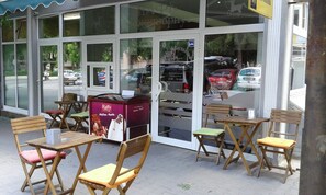 Un área exterior con mesas y sillas de madera, una máquina expendedora y un escaparate de vidrio que refleja coches y árboles.