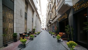 Una calle estrecha flanqueada por macetas con plantas y mesas al aire libre, que lleva a un edificio con un letrero que dice "Bay Window Cafe".