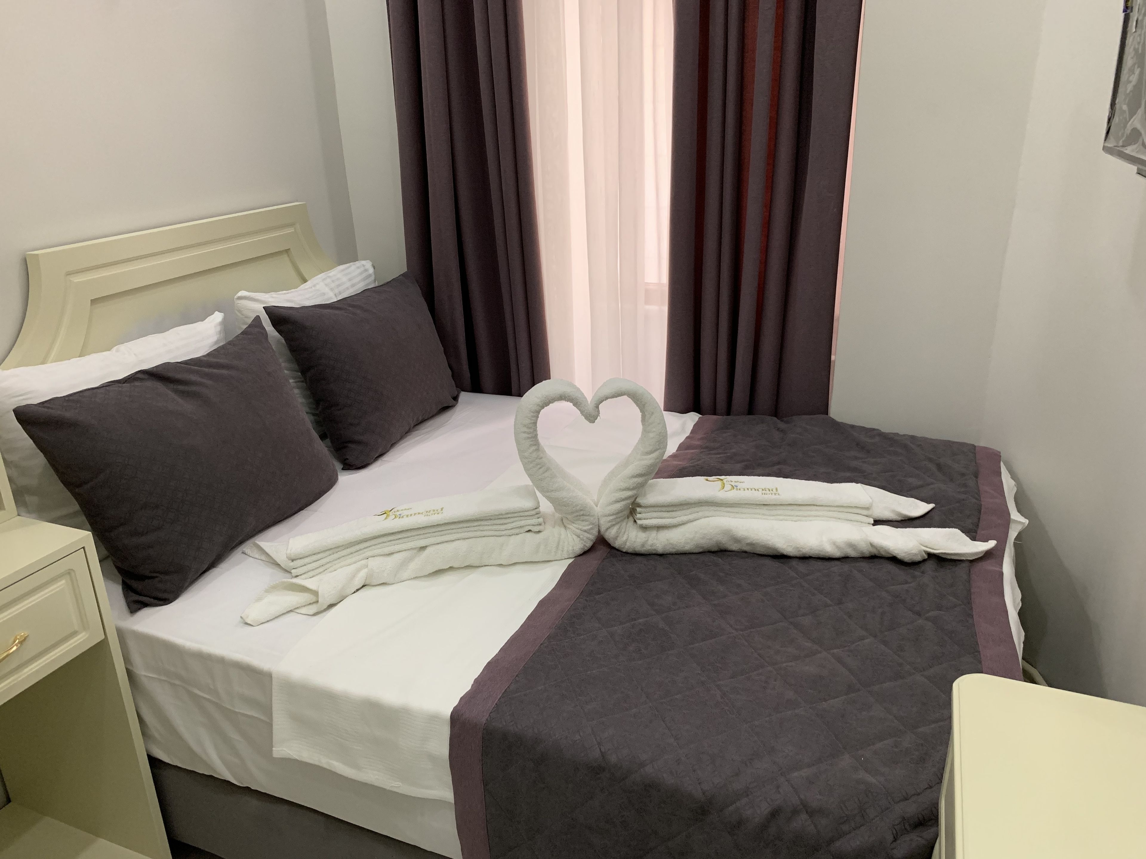 A neatly made bed with a heart-shaped towel arrangement, dark and light pillows, and a grey quilted bedspread.