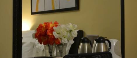 A hotel room with a desk, a vase with flowers, a coffee maker, and a kettle.