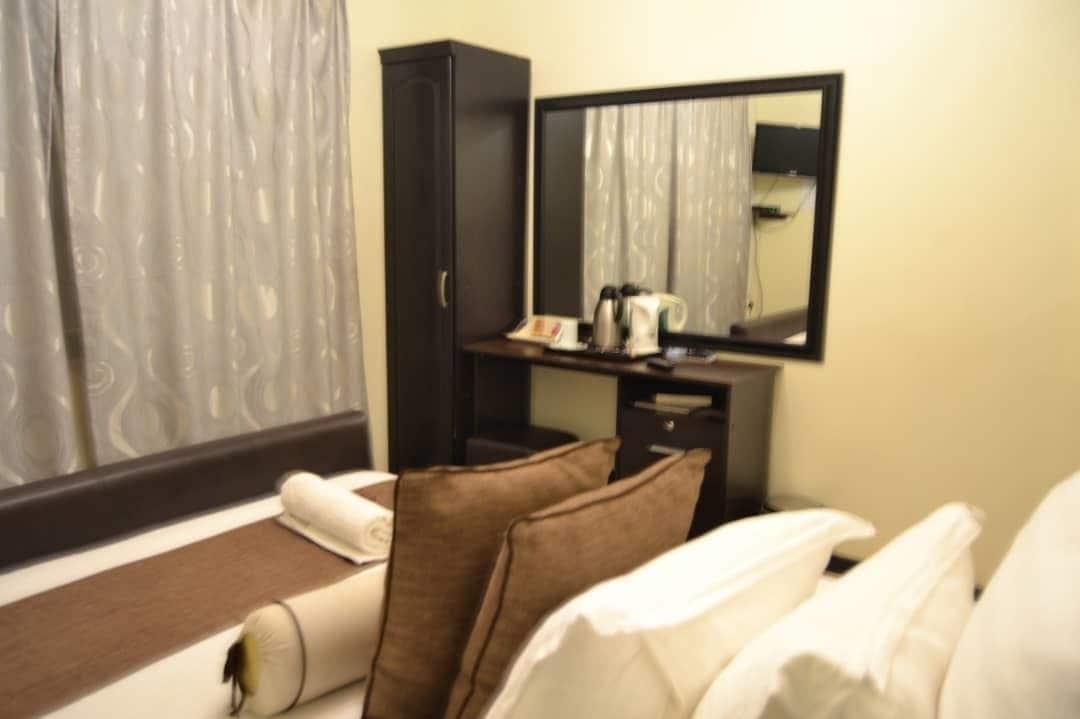 A hotel room with a bed, pillows, a bedside table with a kettle, a mirror, and a wardrobe.